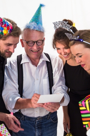 Eine Frau mit Hut pustet Kerzen auf einem Geburtstagskuchen aus. Zwei weitere Personen halten Luftballons in den Händen.
