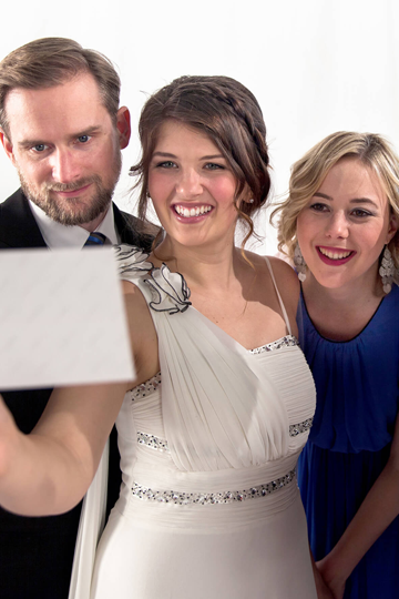 Ein Brautpaar und eine Frau schauen glücklich auf ein Foto.