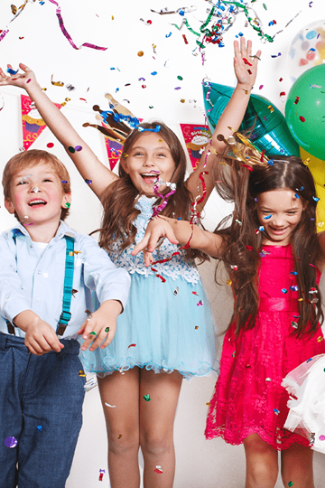 Drei glückliche Kinder mit bunten Luftballons im Hintergrund werfen Konfetti in die Luft.
