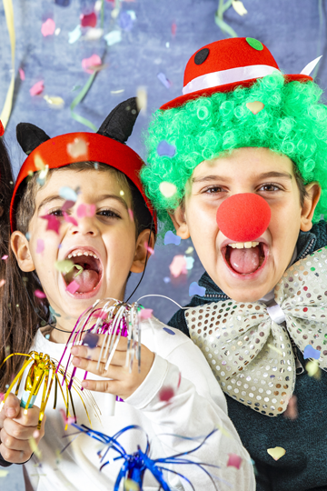 Zwei verkleidete Kinder schauen lachend in die Kamera. Konfetti fliegt durch die Luft.