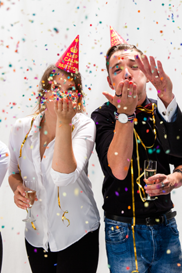 Zwei Personen mit Partyhüten pusten Konfetti von ihrer Hand in die Luft.