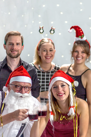 Fünf glückliche Personen mit Weihnachtsrequisiten auf dem Kopf. Zwei Personen stoßen mit Tassen an.